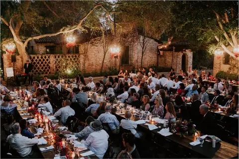 Wedding guests share dinner at long tables beneath trees and hanging lights.
